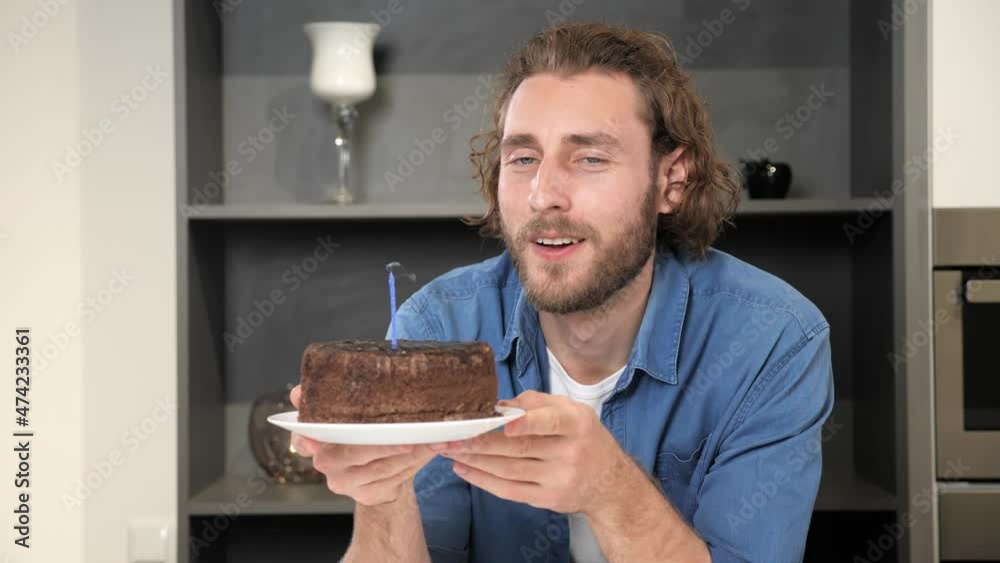 Young happy man celebrating birthday alone. Handsome male bday with party cake and blowing candle. Guy looking satisfied in to camera having nice mood cheerful smile, positive emotion.