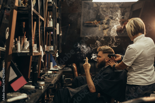 Young bearded man getting haircut by hairdresser and smoking a cigarette