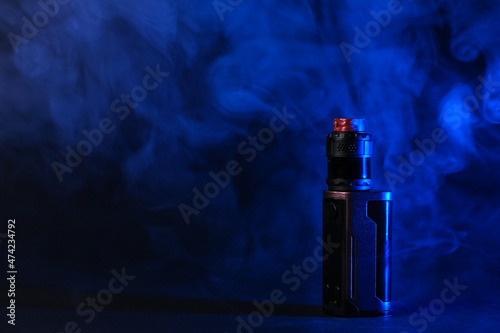 Electronic vaping device mod, atomizer stands in vape smoke and vapor illuminated by blue light.