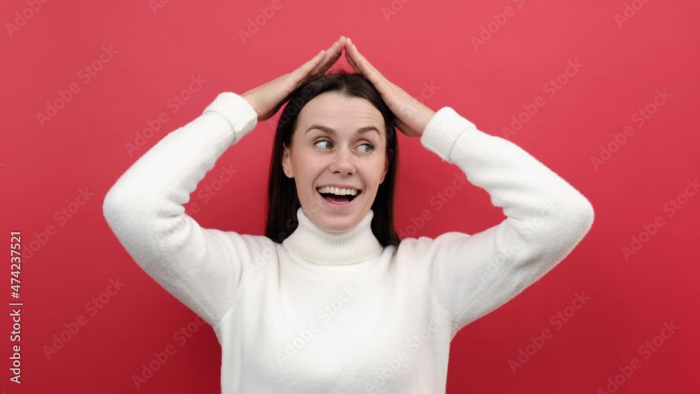 Portrait of positive happy young brunette female standing and raising hands, showing roof gesture, wears cozy knitted sweater, posing isolated over red color background in studio. Dreaming of house Portrait of positive happy young brunette female standing and raising hands, showing roof gesture, wears cozy knitted sweater, posing isolated over red color background in studio. Dreaming of house