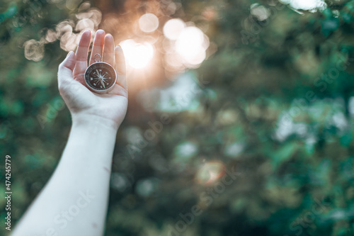 Fototapeta Hand with compass on green blurred background with sun glare at sunset sky