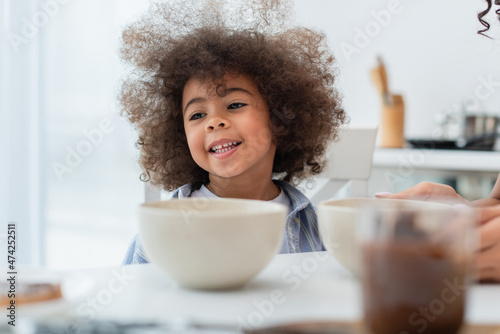 Smiling african american kid sitting near bowls and mom in kitchen.
