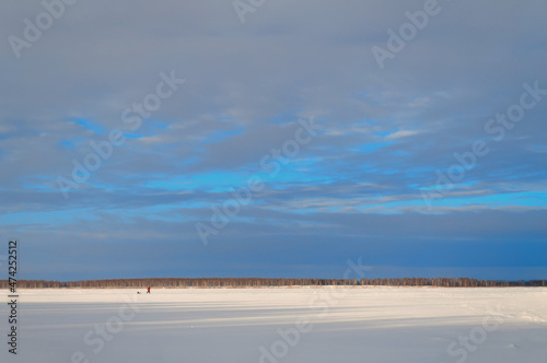 Fototapeta Naklejka Na Ścianę i Meble -  The landscape of a frozen icy lake with an arrow-shaped trail in the snow and a lonely figurine of a man in a red jacket against a blue sky on a sunny day.