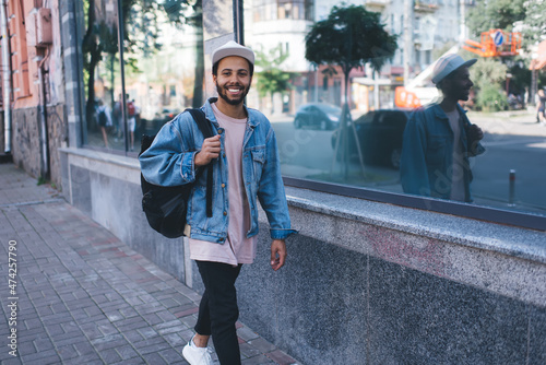 Portrait of trendy dressed Latino student walking at city streets and smiling at camera during free time, cheerful Hispanic tourist with black backpack enjoying travel vacations during spring holidays #474257790