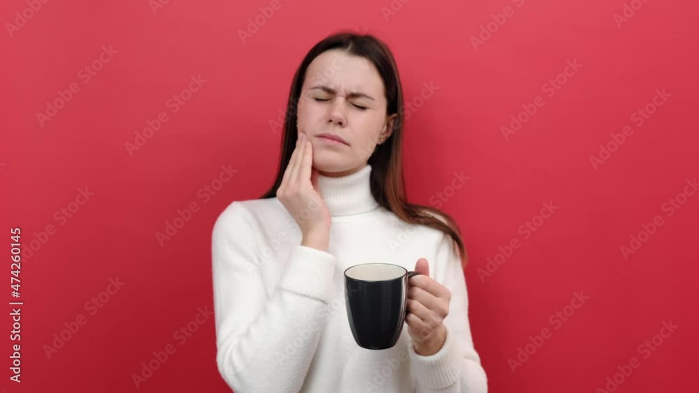 Unhappy young caucasian woman suffering from strong pain after drinking hot or cold beverage from grey cup, wearing white knitted sweater, posing isolated over pastel red color background in studio Unhappy young caucasian woman suffering from strong pain after drinking hot or cold beverage from grey cup, wearing white knitted sweater, posing isolated over pastel red color background in studio