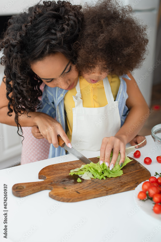 African american woman cutting lettuce near kid in apron in kitchen.
