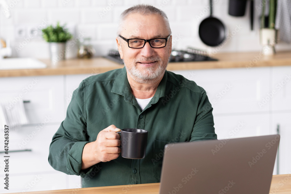 Portrait of happy elderly man with cup of tea and laptop while sitting at table at home