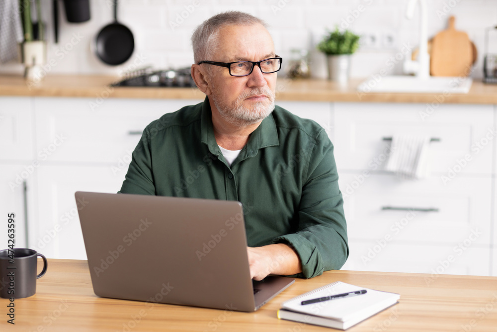 Elderly man working at laptop computer sitting at the table in the home kitchen Elderly man working at laptop computer sitting at the table in the home kitchen