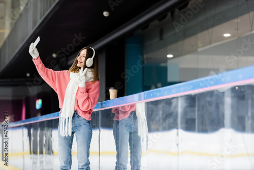 positive young woman taking selfie while showing peace sign near paper cup on ice rink.