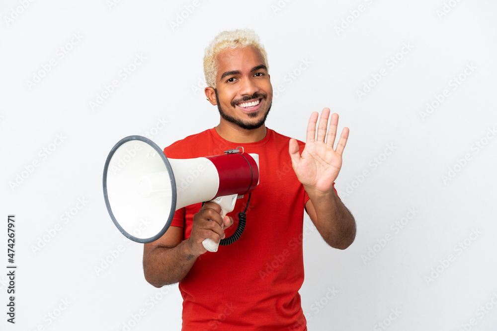 Young Colombian handsome man isolated on white background holding a megaphone and saluting with hand with happy expression Young Colombian handsome man isolated on white background holding a megaphone and saluting with hand with happy expression