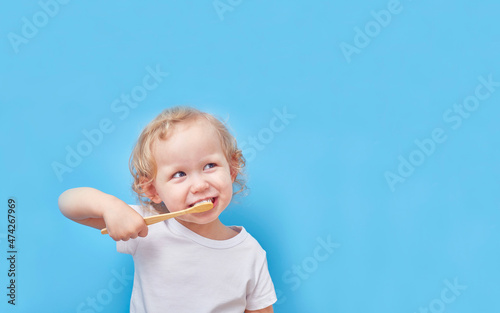 Child brushes his teeth with a wooden bamboo toothbrush on a blue background with a copy space.