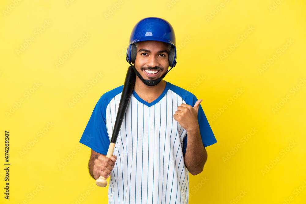 Young Colombian latin man playing baseball isolated on yellow background pointing to the side to present a product