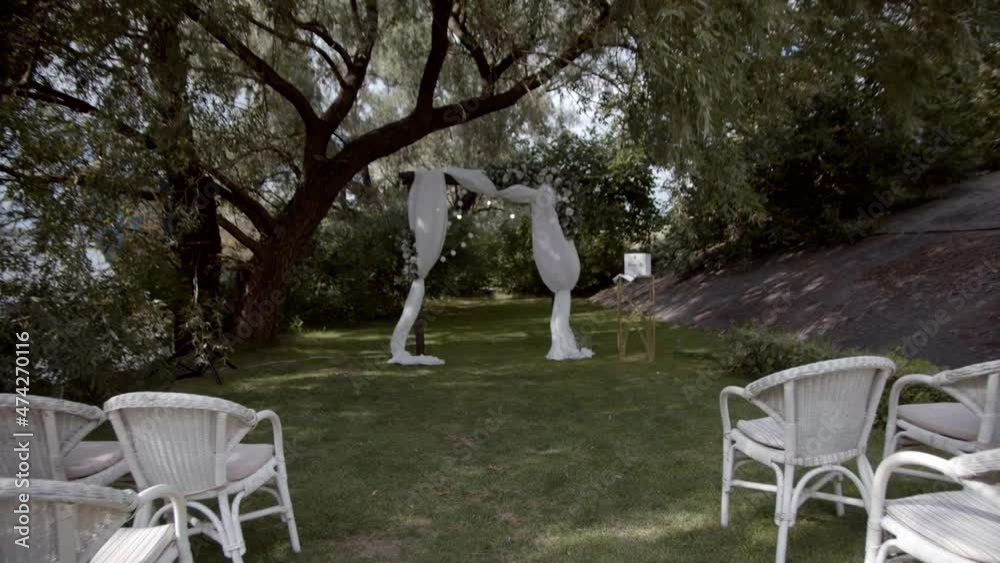 Green area of wedding registration on green lawn there is wedding arch decorated with flowers and white cloth, there are rows of white chairs. Slow motion. Green area of wedding registration on green lawn there is wedding arch decorated with flowers and white cloth, there are rows of white chairs. Slow motion.