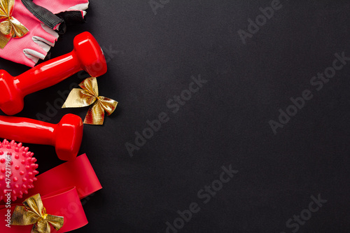 A set of tools for sports for birthday, New year, Christmas. Red dumbbells, fitness bands, gloves, massage ball and golden bows on a black background. Top view, flat lay, copy space, space for text