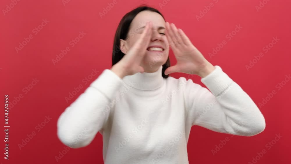 Angry young brunette female 20s keeps arms around wide open mouth, screaming loudly, calling attention to news, wears knitted sweater, posing isolated on red studio background. People emotion concept Angry young brunette female 20s keeps arms around wide open mouth, screaming loudly, calling attention to news, wears knitted sweater, posing isolated on red studio background. People emotion concept