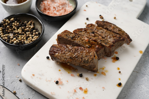 A piece of cooked beef meat steak on white marble board cut in slices, white and black peer, coarse sea salt