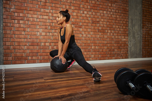 Charming young African athlete in a stylish tight-fitting tracksuit performing side lunges, leaning her hands on a medicine ball