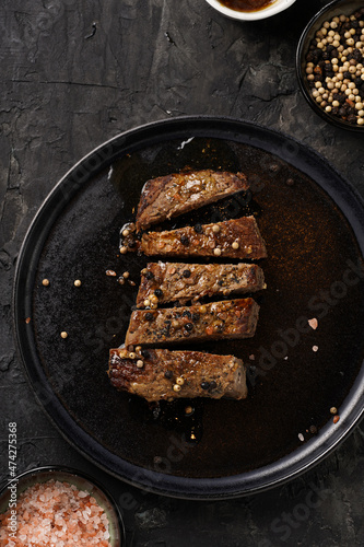 A piece of cooked beef meat steak on black plate cut in slices, white and black peer, coarse sea salt