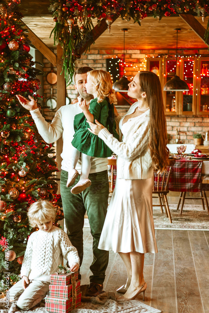 family of four parents pregnant mom and dad and two siblings little boy and girl in stylish clothes in chalet are ready to welcome Christmas and New Year near Christmas tree with gift box presents