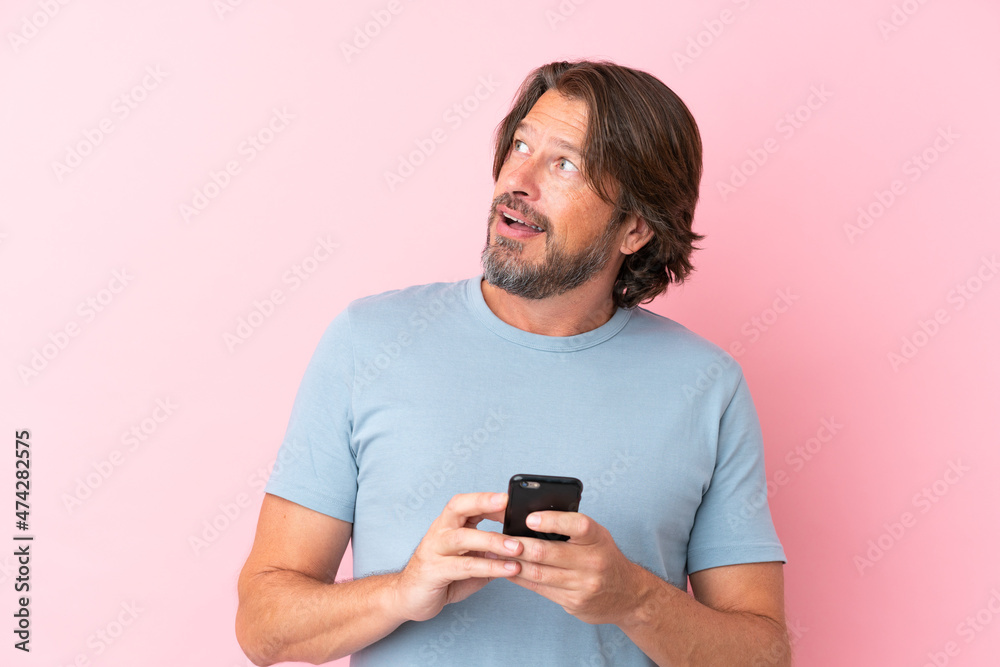 Senior dutch man isolated on pink background using mobile phone and looking up Senior dutch man isolated on pink background using mobile phone and looking up