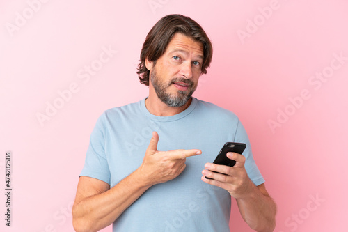 Senior dutch man isolated on pink background using mobile phone and pointing it © luismolinero