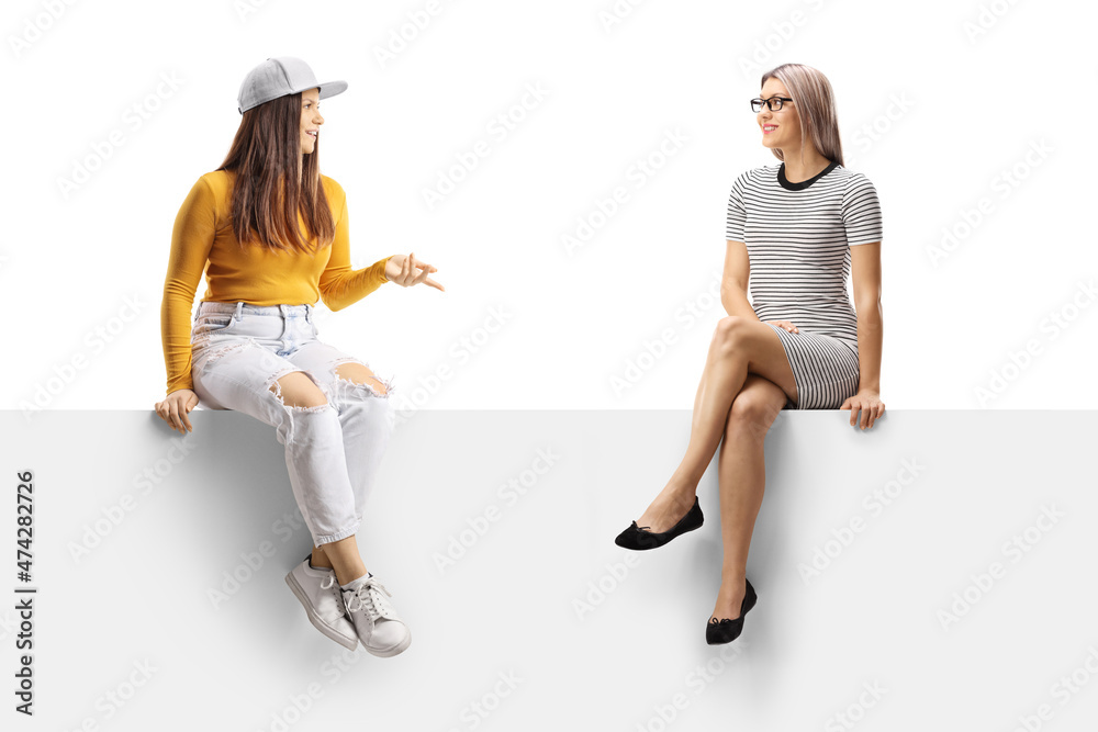 Two young women sitting on a blank panel and having a conversation Two young women sitting on a blank panel and having a conversation