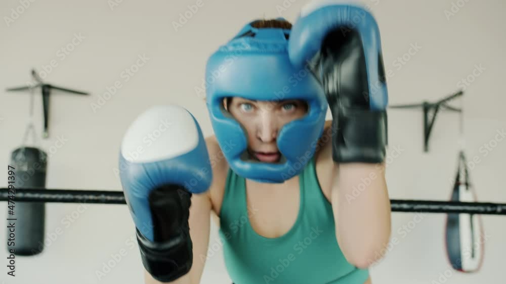 Confident girl boxer wearing protective helmet and gloves is throwing punches looking at camera in gym. Modern sports and lifestyle concept.