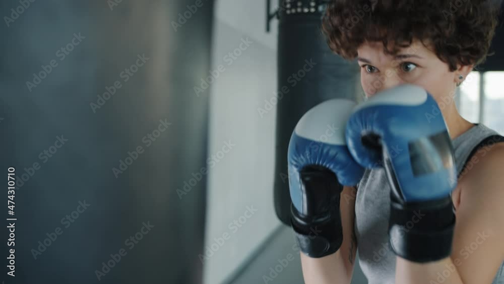Fit young lady is training with punching bag hitting it with gloves exercising in gym alone concentrated on workout. People and sports concept. Fit young lady is training with punching bag hitting it with gloves exercising in gym alone concentrated on workout. People and sports concept.