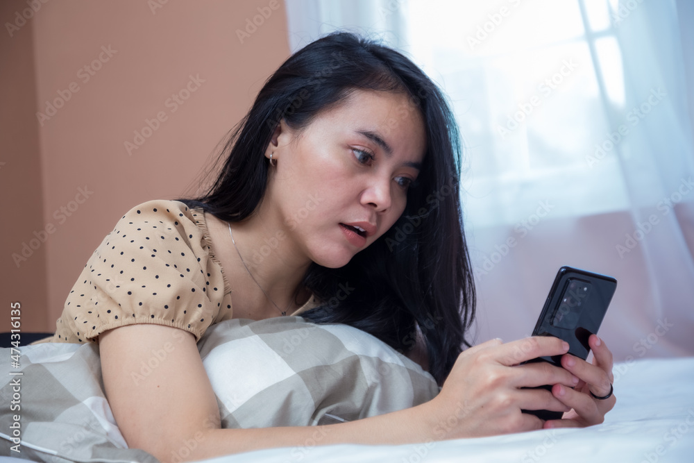 Woman lying on bed while holding cell phone with weird facial expression Woman lying on bed while holding cell phone with weird facial expression