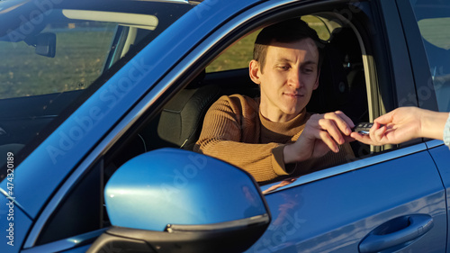 Woman hands over remote control key of bright blue foreign car to young man driver sitting in automobile in sweater with brunet hair and smile photo