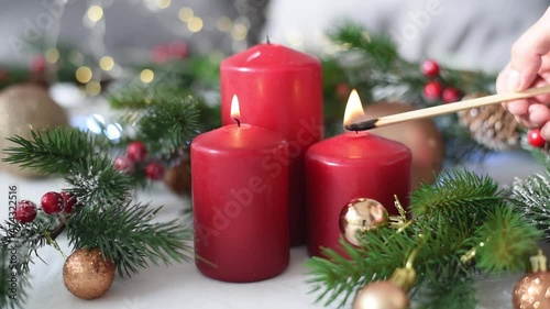 Hand of woman holds burning stick and light up three red candles. Christmas decoration with spruce branches, cones, golden balls and garland on blurry background. Side view. new year mood