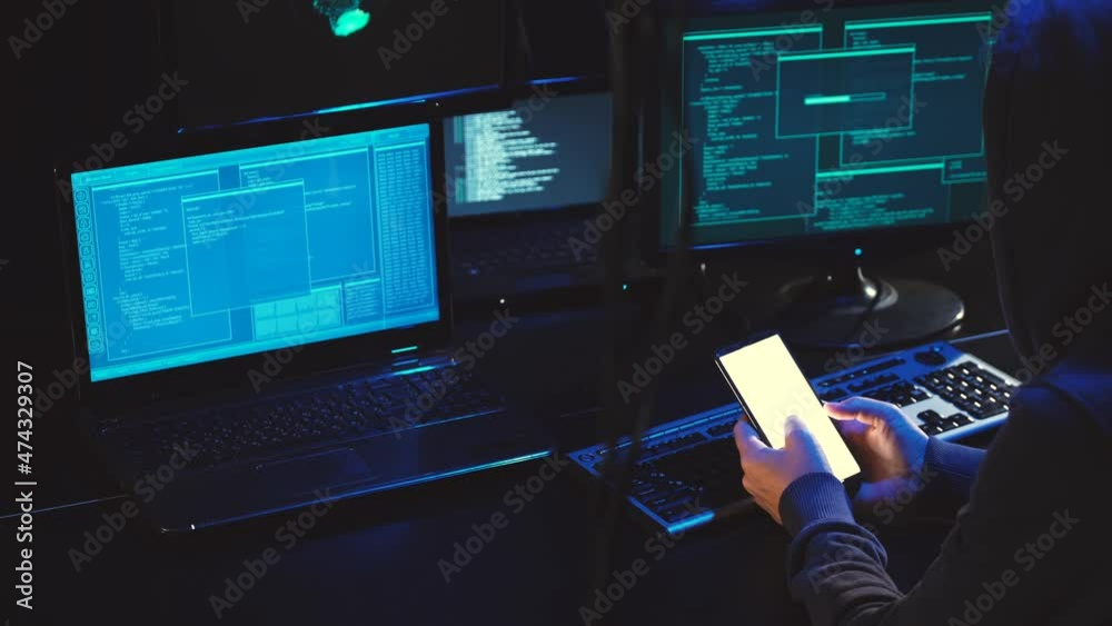 Overhead hacker in hood working at laptop and mobile phone typing text in dark room, An anonymous hacker uses malware with mobile phone to hack password, personal data steals money from bank. cyber