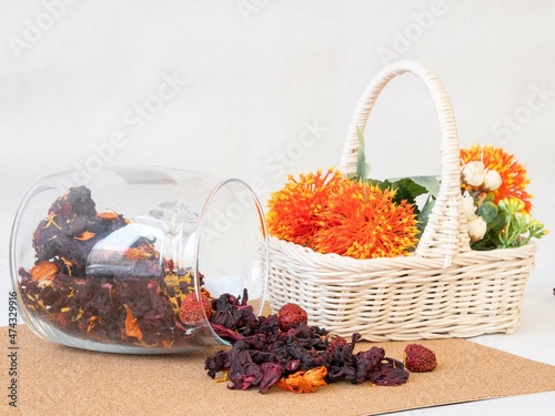 Flavored red tea made from hibiscus rose petals with dry berries is poured from a glass jar onto a cork napkin next to a basket of flowers. Tea business products. Tea blog content.