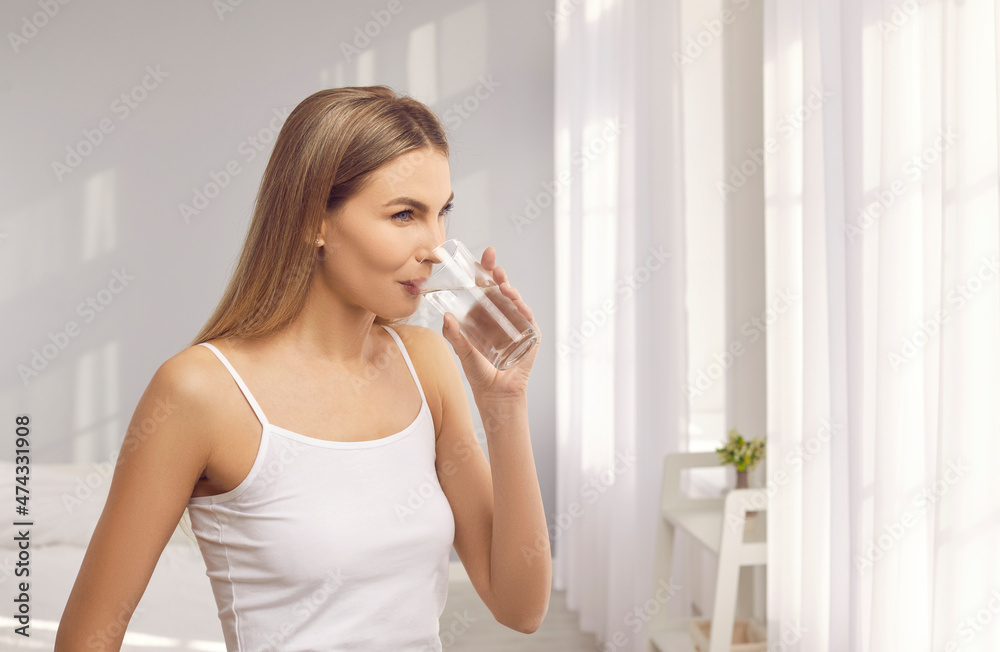 Portrait of pretty woman standing by bedroom window and enjoying glass of purified water to remind you that drinking enough water daily is a good habit. Healthy lifestyle and staying hydrated concept Portrait of pretty woman standing by bedroom window and enjoying glass of purified water to remind you that drinking enough water daily is a good habit. Healthy lifestyle and staying hydrated concept