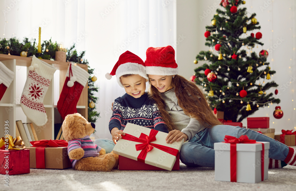 Magic time. Cute happy kids boy and girl in Santa hats open box with Christmas presents on festive morning. Sister and brother open gift box sitting on warm floor in room with Christmas decorations.