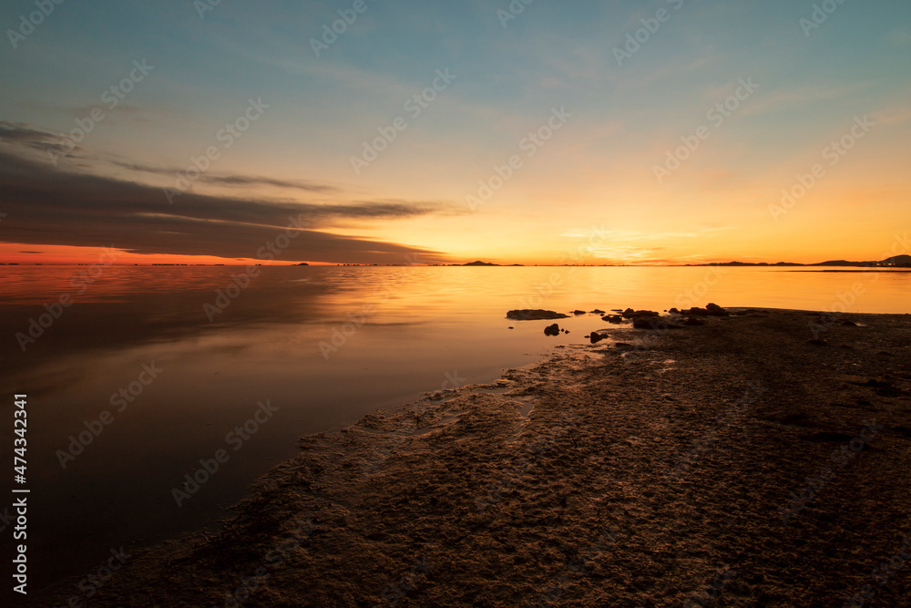 Photograph of a sunrise in the Mar Menor, in the region of Murcia, Spain. The sea is calm and there are orange clouds that are reflected on the shore of the beach
