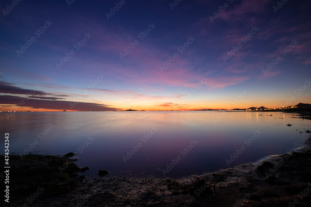 Photograph of a sunrise in the Mar Menor, in the region of Murcia, Spain. The sea is calm and there are pink clouds that offer a beautiful reflection in the water