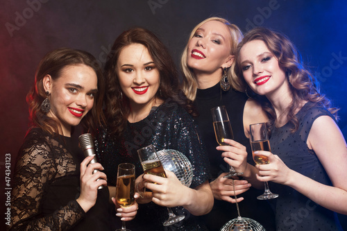 Group of partying women clinking flutes with sparkling wine and holding disco balls and microphone.