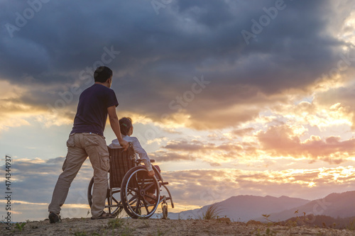 Woman Sitting in wheelchair and her care helper walking on mountain at sunset background. photo