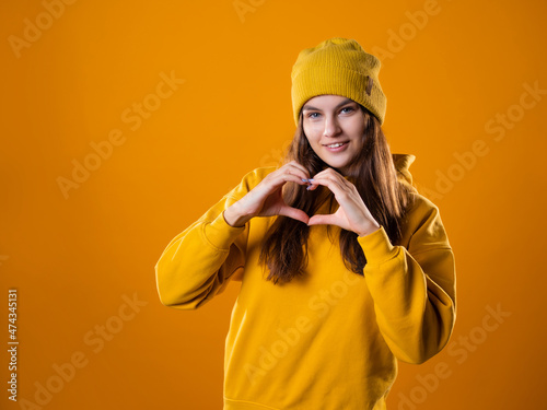 Stylish cheerful young brunette in a yellow hoodie and hats shows a heart sign gesture, on a yellow background