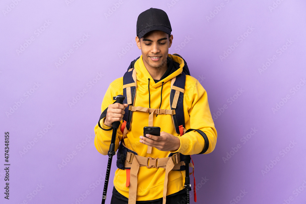 African American man with backpack and trekking poles over isolated background sending a message with the mobile African American man with backpack and trekking poles over isolated background sending a message with the mobile