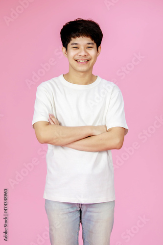 Portrait studio shot of Asian young handsome happy teenager short hair male model in casual outfit white shirt and denim jeans standing alone smiling crossed arms look at camera on pink background