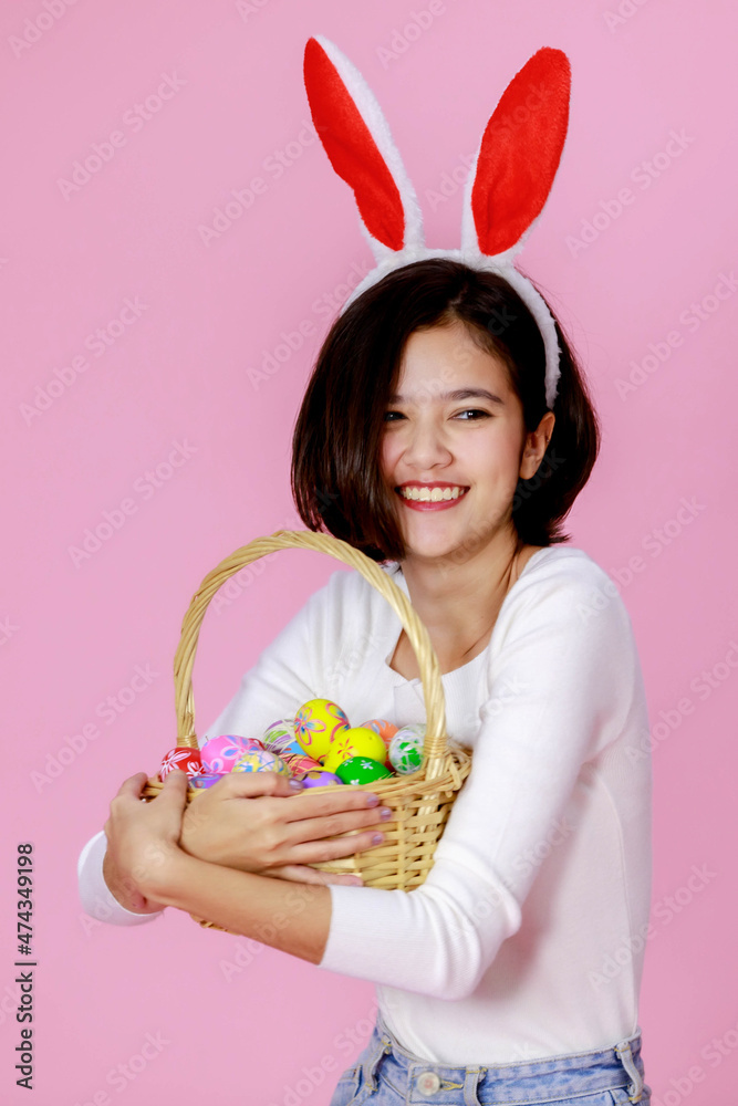 Asian young happy pretty short hair teenager girl model with bunny ears headband standing holding basket of traditional festival colorful painted easter eggs on sticks on pink background Asian young happy pretty short hair teenager girl model with bunny ears headband standing holding basket of traditional festival colorful painted easter eggs on sticks on pink background