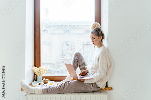 Photography Young blond woman freelancer in cozy knitted white sweater sitting on windowsill with laptop computer working from home