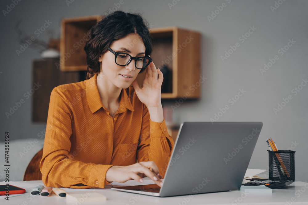 ocused Spanish woman in glasses sits at desk and typing on laptop during remote work at home ocused Spanish woman in glasses sits at desk and typing on laptop during remote work at home