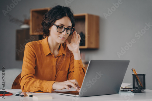 ocused Spanish woman in glasses sits at desk and typing on laptop during remote work at home