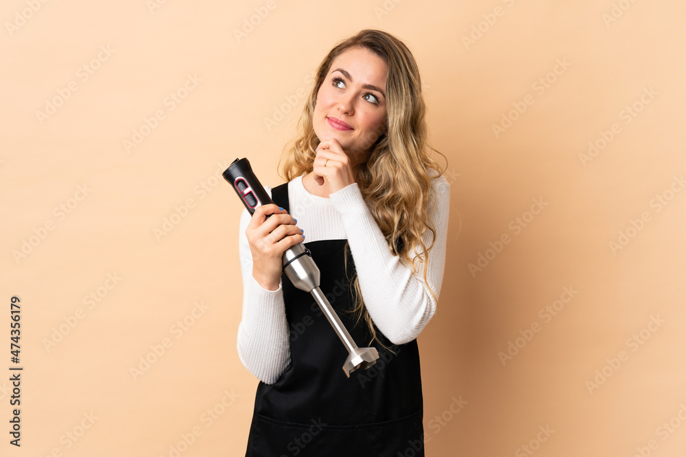 Young brazilian woman using hand blender isolated on beige background and looking up Young brazilian woman using hand blender isolated on beige background and looking up
