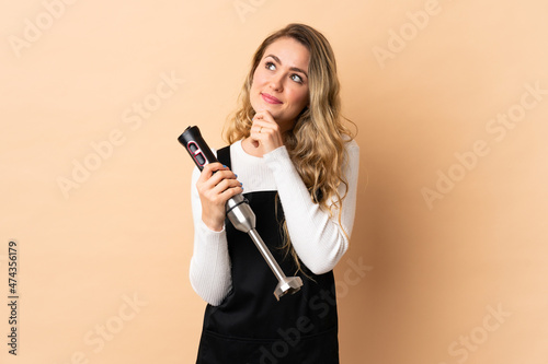 Young brazilian woman using hand blender isolated on beige background and looking up