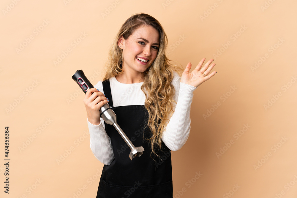 Young brazilian woman using hand blender isolated on beige background saluting with hand with happy expression