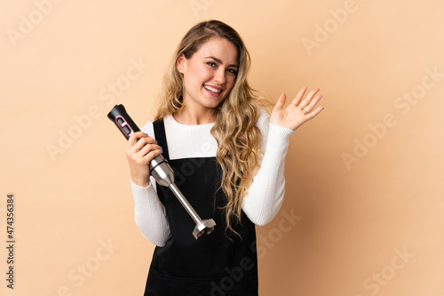 Young brazilian woman using hand blender isolated on beige background saluting with hand with happy expression